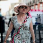 A fashionable woman in a floral dress and hat walking confidently on a sunny city street.