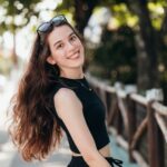A young woman with long hair smiles while standing outdoors in a sunny setting.
